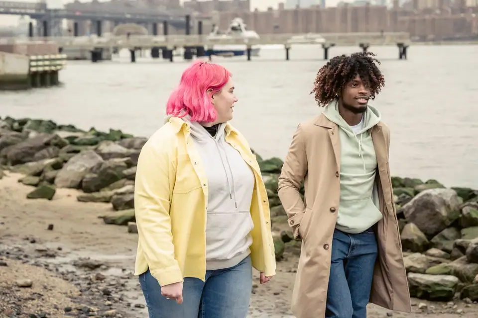 A couple walking on a stony riverbank, both in hoodies under open jackets