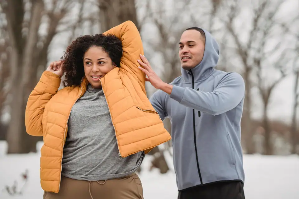 A woman in an orange puffer jacket and a man in a gray zip hoodie laughing in a snowy park